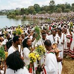 Thousands of people in traditional Oromo clothing gather at the edge of a lake during the Irreecha festival in Addis Ababa, holding green grass and yellow flowers.
