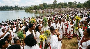 Thousands of people in traditional Oromo clothing gather at the edge of a lake during the Irreecha festival in Addis Ababa, holding green grass and yellow flowers.